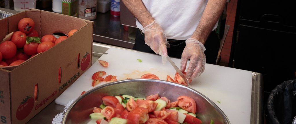 A chef cuts a tomato into wedges to add to a large bowl of cucumbers and tomatoes. A large box full of tomatoes sits nearby.
