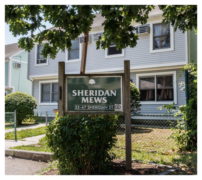 The Sheridan Mews sign sits near a shrub and in front of the first building. The building's windows are open, letting in fresh air.
