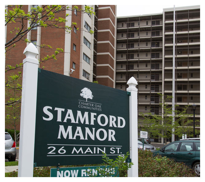 The large Stamford Manor sign sits in the parking lot with the impressive, eleven-story building towering behind it.