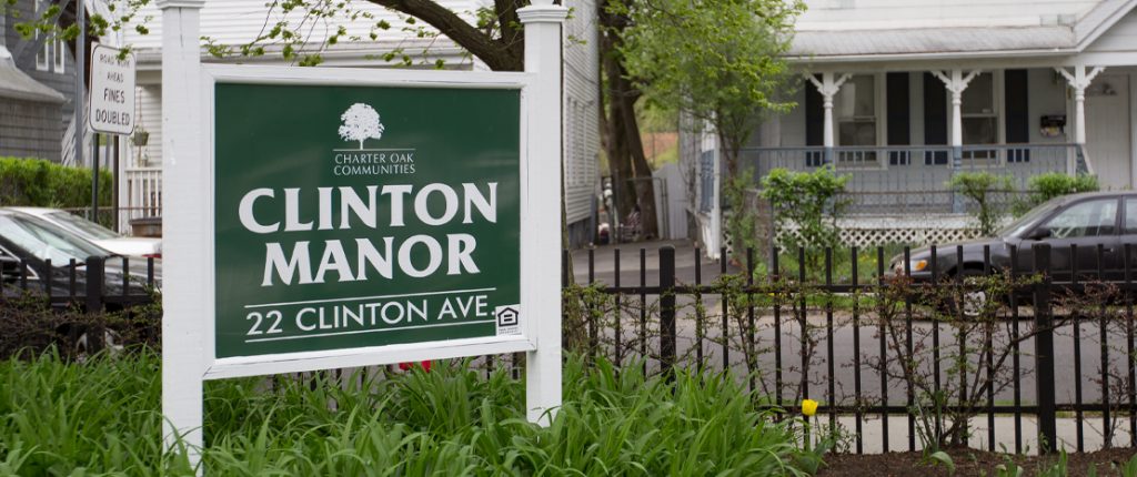The quaint sign for Clinton Manor sits among plants and flowers and reads Clinton Manor, 22 Clinton Avenue.