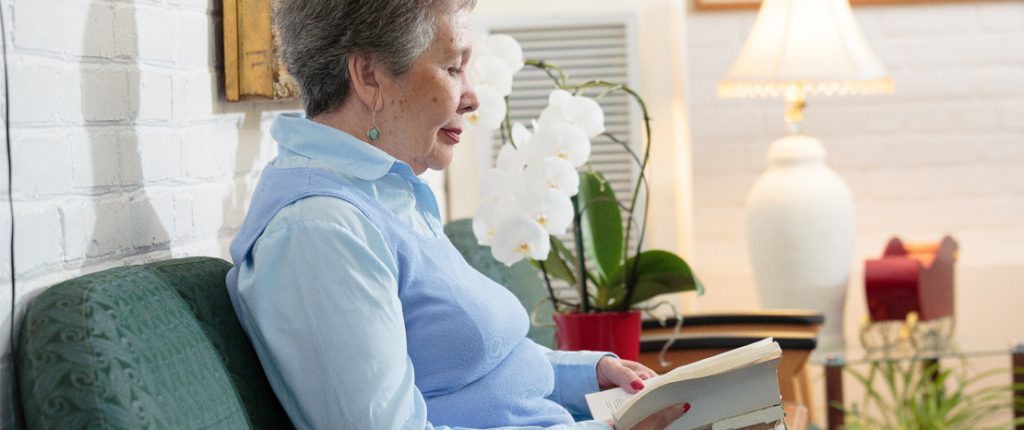 A senior resident sits in a comfortable-looking chair reading a book. Orchids sit on a nearby table with a bright lamp.