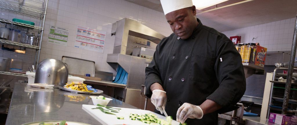The on-site chef, dressed in his black chef coat and tall hat, cuts up cucumbers in the clean and organized facility kitchen.