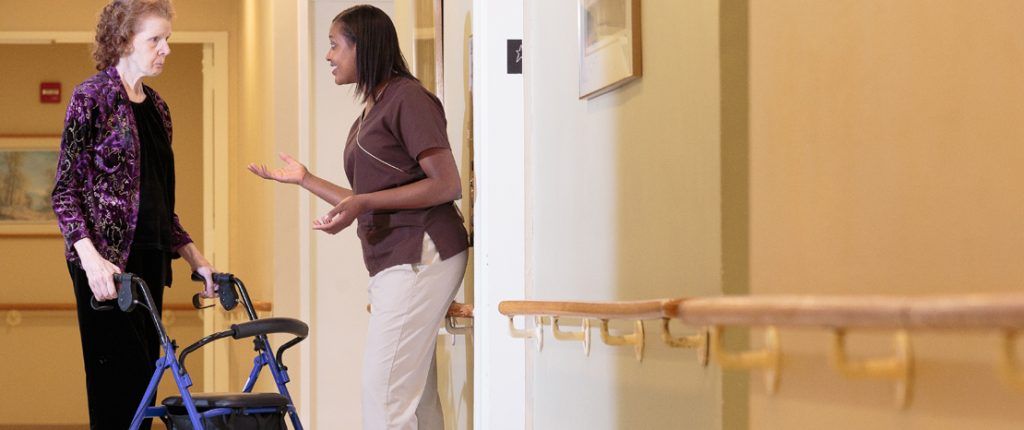 An on-site registered nurse engages a resident using a rollator in conversation. Support railings line the walls.